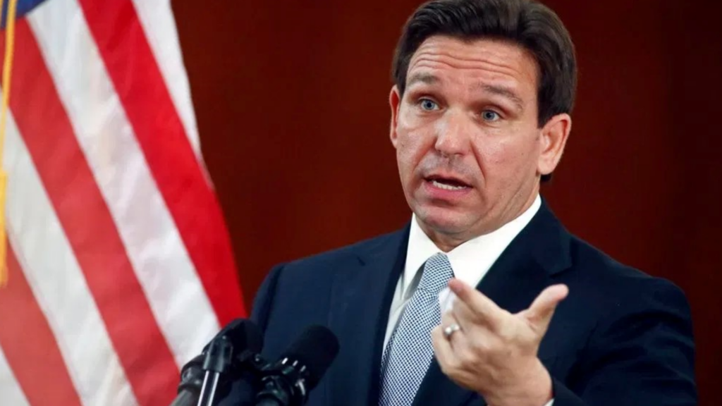 A man in a dark suit and striped tie speaks into microphones, gesturing with his hand. An American flag is visible in the background.