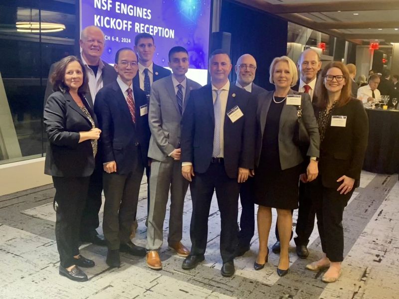 A group of ten people in business attire stand together, smiling for a photo at the NSF Engines Kickoff Reception, as indicated by the banner in the background. They are indoors, on a carpeted floor.