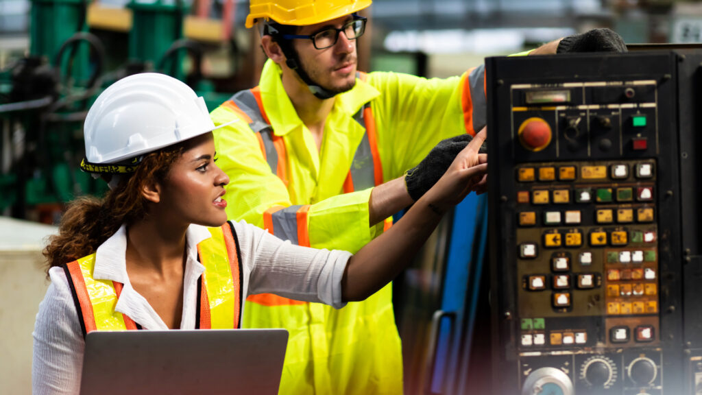A woman and a man wearing safety helmets and reflective vests operate an industrial control panel. The woman holds a laptop while the man points to buttons on the machine in a factory setting.