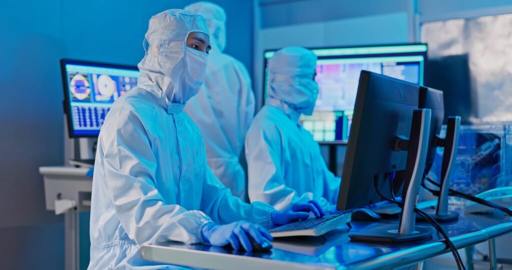 Two scientists in full protective suits work at computer stations in a high-tech laboratory, with multiple monitors displaying data in the background. The setting is brightly lit with a blue tone.