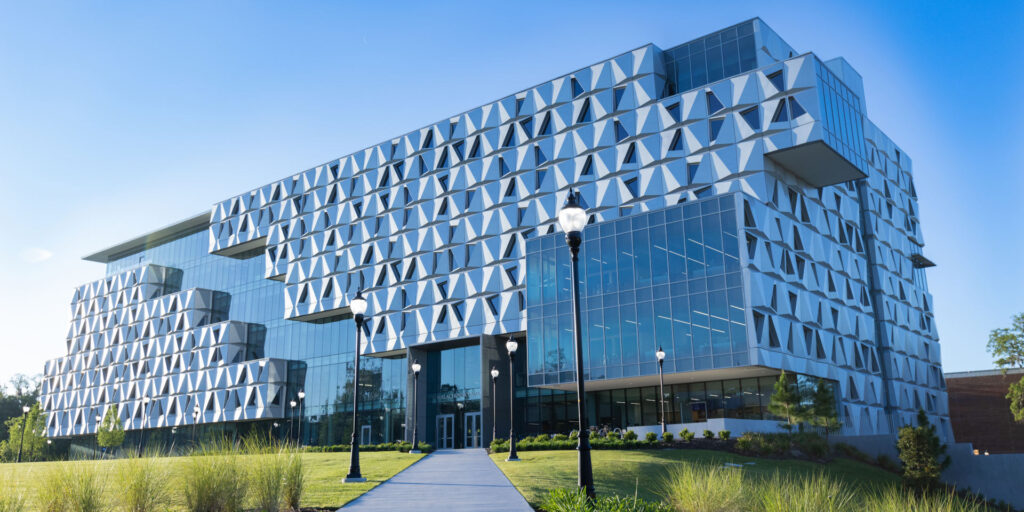 Modern glass building with a unique geometric facade, featuring triangular patterns. The structure is surrounded by green landscaping, including grass and small bushes, and lit by black street lamps under a clear blue sky.
