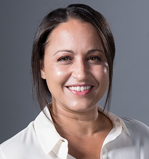A woman with straight dark hair, parted to the side, smiles while wearing a white collared shirt. The background is plain and gray.